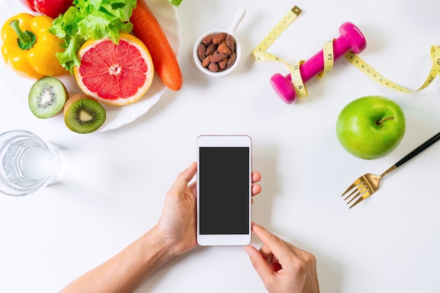 Mãos segurando um telefone móvel no fundo da mesa branca. conceito de alimentação, dieta e exercícios limpos