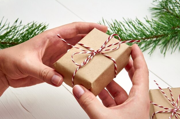 Mãos segurando uma caixa de presente de Natal sobre uma mesa branca com galhos de pinheiros