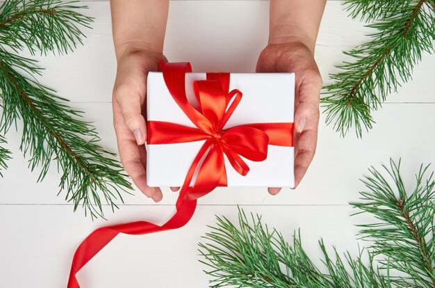 Mãos segurando uma caixa de presente de natal sobre uma mesa branca com galhos de pinheiros