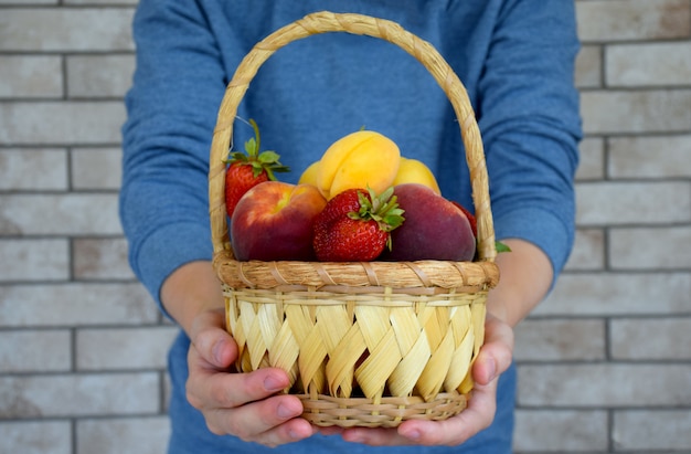 Foto mãos segurando uma cesta de vime cheia de frutas orgânicas maduras no fundo da parede de tijolo