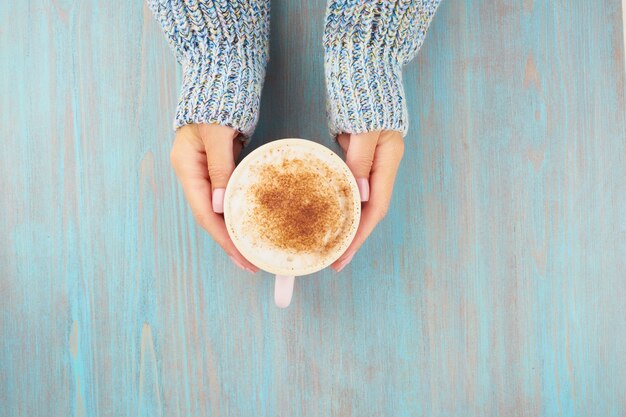 Mãos segurando uma xícara de chocolate quente, mesa de madeira azul, blusa aconchegante azul, linda manicure rosa