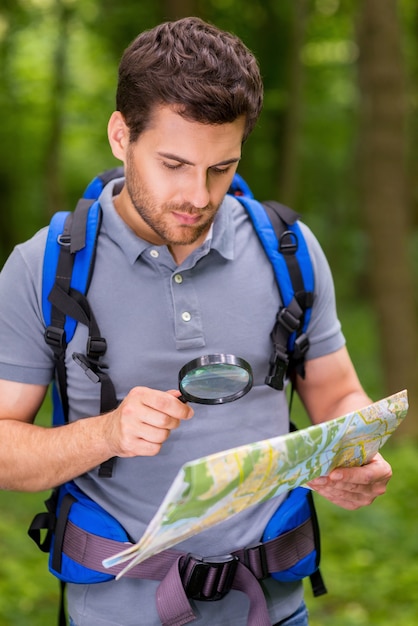 Mapa de examen de turista confiado. Hombre joven seguro con mochila examinando el mapa con lupa mientras está de pie en la naturaleza