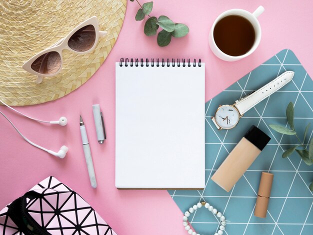 Maqueta de bloc de notas en blanco. Escritorio rosa femenino plano con cosmético, sombrero de paja, té y bloc de notas abierto. Copie el espacio.