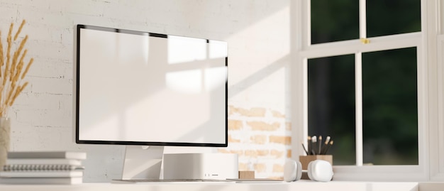 Maqueta de computadora de pantalla blanca, auriculares y artículos de papelería en una mesa blanca contra una pared de ladrillo blanco