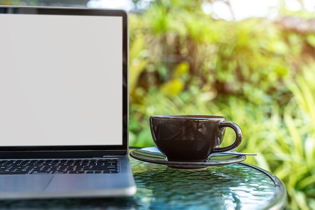 Maqueta de computadora portátil con pantalla vacía con taza de café y teléfono inteligente en la mesa de la cafetería al aire libre fondo Pantalla blanca