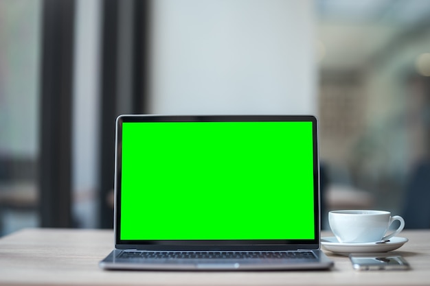 Maqueta de computadora portátil con pantalla vacía con taza de café y teléfono inteligente en la mesa del fondo de la cafetería, pantalla verde