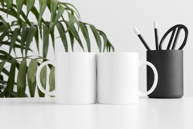 Foto maqueta de dos tazas con accesorios de espacio de trabajo en una mesa blanca y una planta de palma