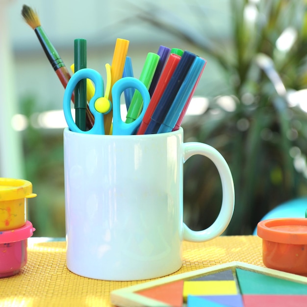 Maqueta del espacio vacío de la taza de té blanco para el logotipo o imprimir una foto al aire libre de la taza blanca vacía en el escritorio de trabajo de arte con lupa de rotuladores de colores y tangram