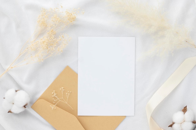 Maqueta de invitación de tarjeta de felicitación en blanco 5x7 en sobre con flores secas y cinta sobre fondo de tela blanca maqueta plana laicoxA