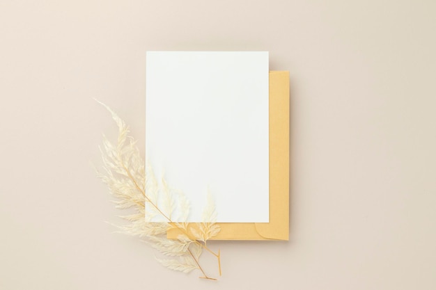 Maqueta de invitación de tarjeta de felicitación en blanco 5x7 en sobre marrón con flores secas sobre fondo beige maqueta plana