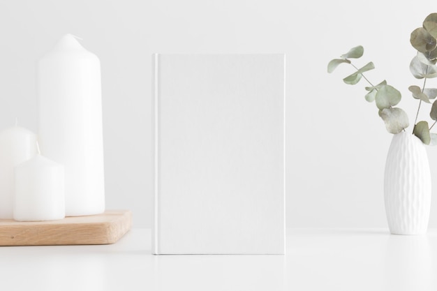 Maqueta de libro blanco con un eucalipto en una olla y velas sobre una mesa blanca