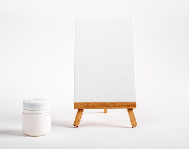 Maqueta de lienzo vertical sobre caballete de madera, tarro de pintura en blanco blanco, herramientas creativas minimalistas, suministros de dibujo para promoción publicitaria