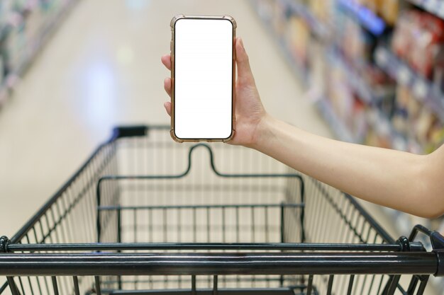 Maqueta, manos sosteniendo un teléfono móvil de pantalla blanca en blanco con carrito de compras en el supermercado