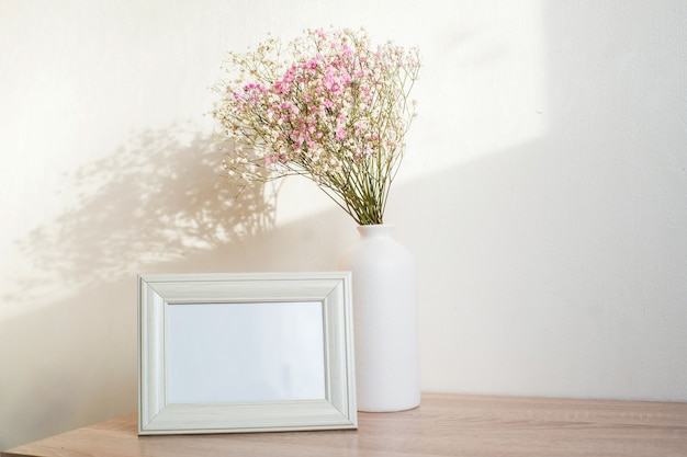 Maqueta de marco blanco horizontal en banco de madera vintage, mesa. Jarrón de cerámica blanca moderna gypsophila. Fondo de pared blanca.