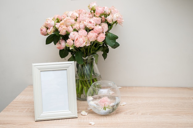 Maqueta de marco de imagen blanco retrato en mesa de madera Jarrón de cristal moderno con rosas Interior escandinavo de pared blanca