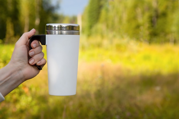 Maqueta de una mujer sosteniendo una taza de viaje blanca junto a un bosque. Taza vacía maqueta para promoción de diseño.