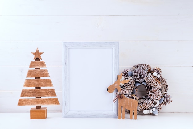 Maqueta de Navidad con marco blanco, corona de conos, árbol de madera y ciervos en madera blanca