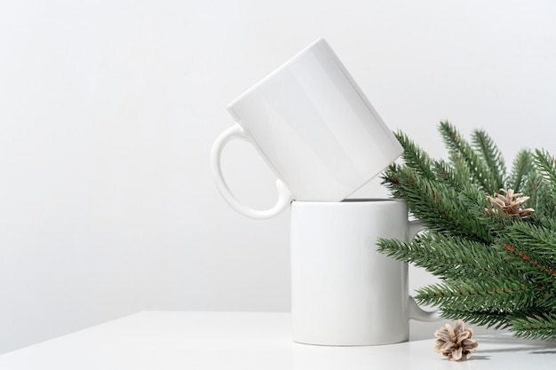 Maqueta de Navidad tazas de té vacías blancas sobre una mesa blanca y ramas de una corona de árbol de Navidad.