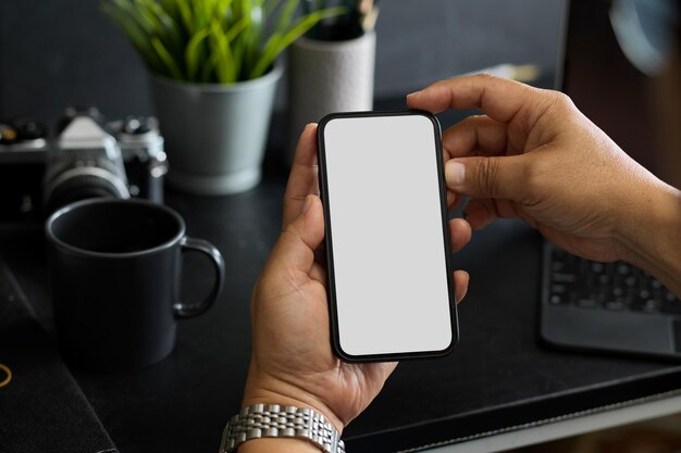 Una maqueta de pantalla en blanco de un teléfono inteligente moderno en un hombre entrega un espacio de trabajo oscuro moderno. de cerca