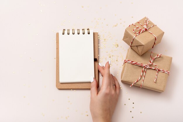 Maqueta de papel en blanco de cuaderno de mano femenina, confeti de estrellas doradas, cajas de regalo sobre fondo beige. Endecha plana, vista superior, espacio de copia, minimalista. Composición de Navidad año nuevo.