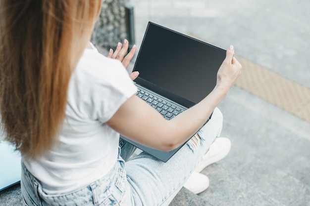Maqueta de portátil. La chica joven estudiante usa una computadora portátil con pantalla blanca, sentada en las escaleras y mirando la pantalla del monitor negro vacío cerca del edificio de oficinas. Copie el espacio para el diseño