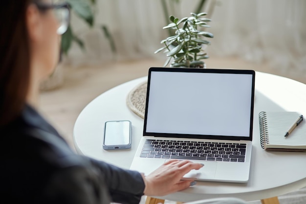 Maqueta portátil de pantalla blanca y mujer de teléfono móvil usando la computadora sentada en la mesa en la vista posterior de la casa