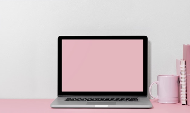 Maqueta de portátil con pantalla en blanco con taza y libro sobre la mesa, diseño de color rosa.