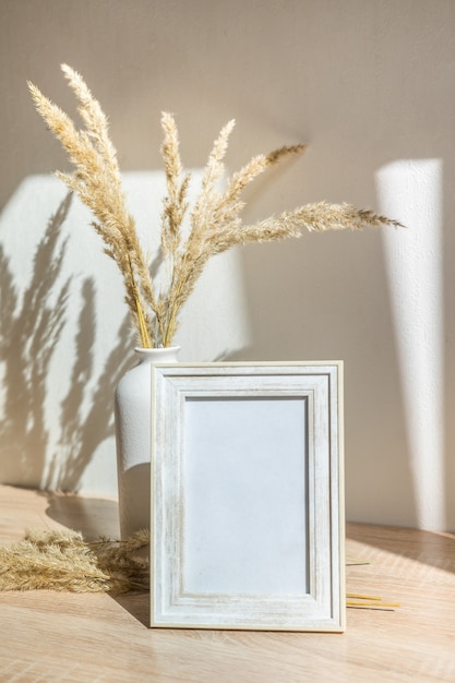 Maqueta de retrato de marco de imagen blanco sobre mesa de madera Jarrón de cerámica moderno con flores secas
