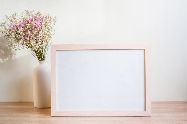 Maqueta de retrato de marco de imagen blanco sobre mesa de madera. Jarrón de cerámica moderno con gypsophila.