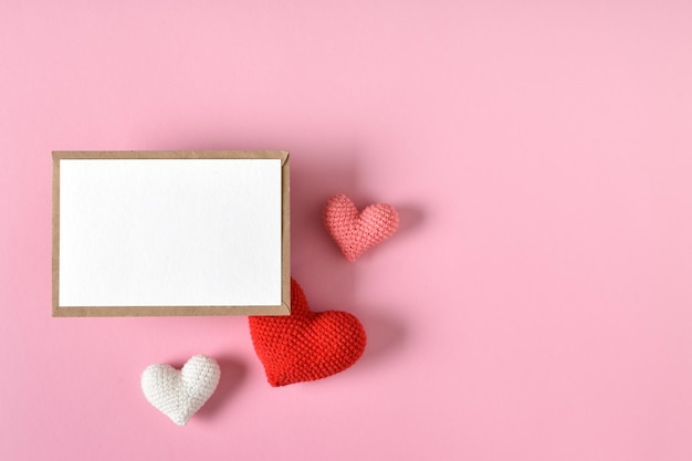 Una maqueta de un sobre kraft con una hoja en blanco y corazones tejidos Tarjeta de felicitación del Día de San Valentín con corazones sobre un fondo rosa