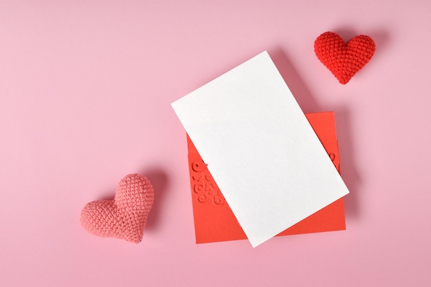 Una maqueta de un sobre rojo con una hoja en blanco y dos corazones de punto sobre un fondo rosa