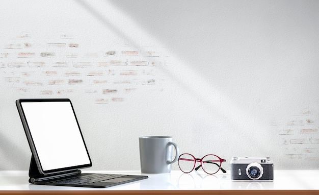 Maqueta de tableta de pantalla en blanco con teclado mágico, cámara vieja, taza y vasos en la mesa de madera y pared de ladrillo.