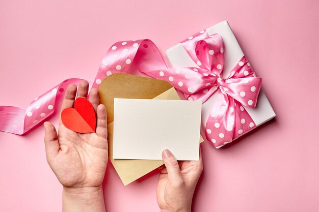 Maqueta de tarjeta de felicitación con caja de regalo con lazo en manos femeninas en tarjeta de felicitación en blanco rosa