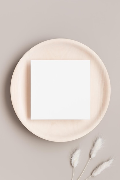 Maqueta de tarjeta de invitación cuadrada en un plato de madera con decoración de lagurus