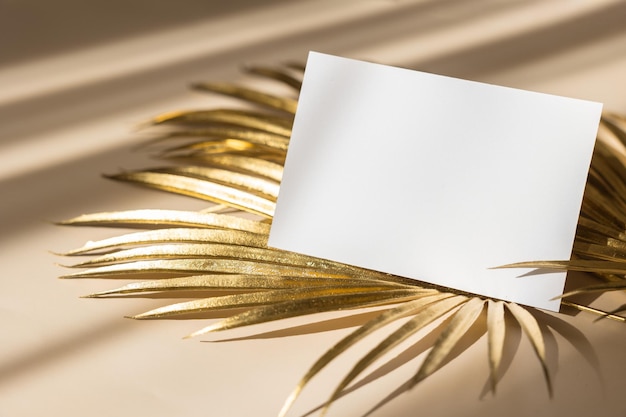 Maqueta de tarjeta de invitación con hojas de palma doradas sobre fondo beige pastel Vista superior espacio de copia plana Plantilla en blanco de maqueta de papel blanco para marca y publicidad