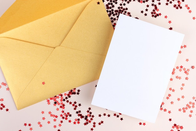 Maqueta de tarjeta de invitación con sobre dorado y papel blanco vacío y confeti en forma de estrella roja