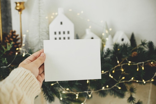 Maqueta de tarjeta de Navidad Mano sosteniendo una tarjeta de felicitación vacía en el fondo de elegantes casas navideñas ramas de abeto con luces doradas y decoraciones de árboles Espacio para texto Plantilla de saludos de temporada