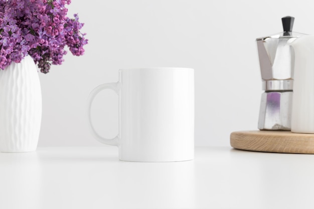 Maqueta de taza blanca con una moka y un ramo de lilas en un jarrón sobre una mesa blanca