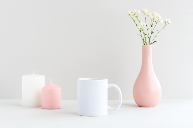 Maqueta de taza blanca con velas, jarrón rosa y rama de gypsophila sobre una mesa