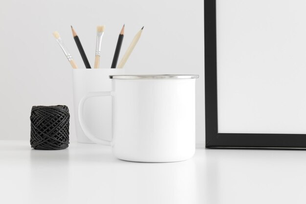 Maqueta de taza de esmalte con accesorios de espacio de trabajo en una mesa blanca