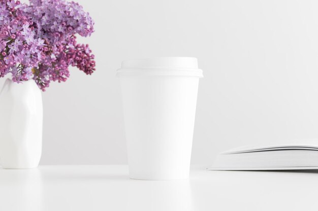 Maqueta de taza de papel de café con un libro y un ramo de lilas en un jarrón sobre una mesa blanca