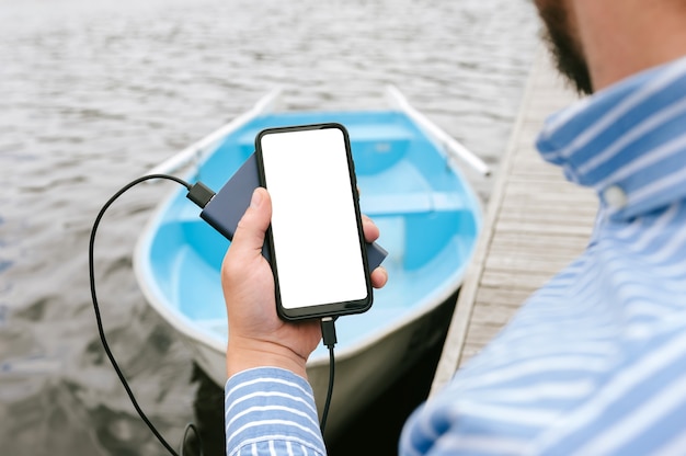 Maqueta de un teléfono inteligente con carga desde el banco de energía en la mano de un hombre. En el contexto de un barco en el agua en el muelle de madera.