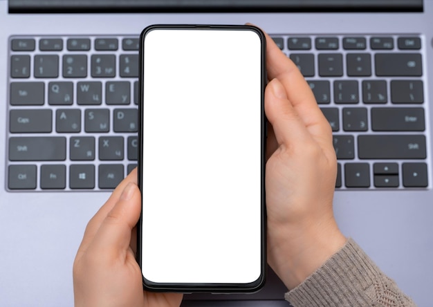 Maqueta de teléfono móvil Imagen de maqueta de vista superior de manos sosteniendo un teléfono móvil de pantalla blanca en blanco con una computadora portátil vista de cerca