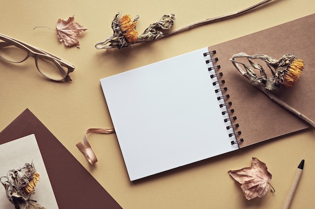 Maquete com caderno de álbum em branco com fichário em espiral óculos de leitura flores secas de girassol postura plana em camadas de papel dourado e off white sombras naturais da luz solar materiais reciclados de baixo impacto