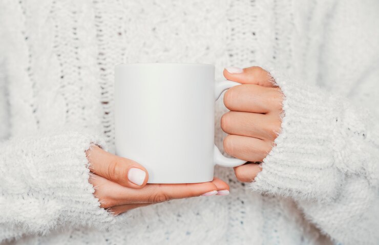 Maquete da caneca branca. Menina está segurando um copo de cerâmica branca de 11 onças nas mãos, usa uma camisola de malha aconchegante. Vista frontal, copie o espaço.