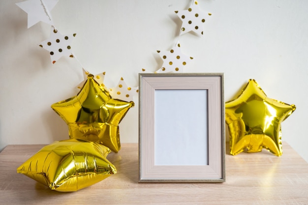Maquete da moldura branca do retrato na mesa de madeira Balões dourados Conceito de aniversário Fundo da parede branca Interior escandinavo Vertical