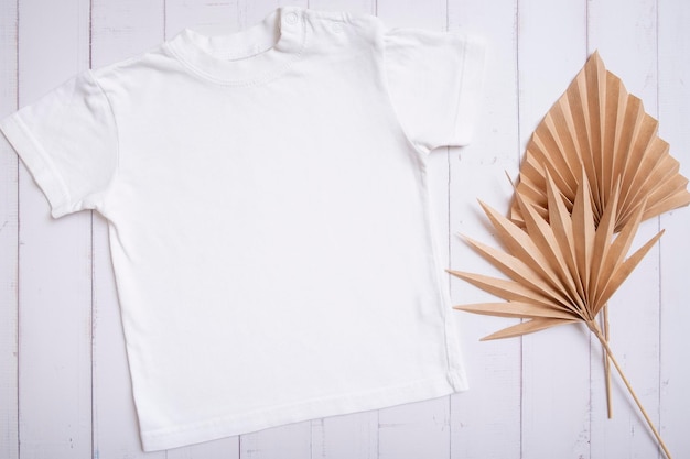 Maquete de camiseta infantil branca para texto de logotipo ou design em fundo de madeira com vista superior de folhas de palmeira