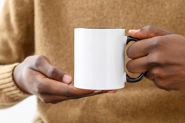 Maquete de caneca de cerâmica fecha as mãos afroamericanas segurando um copo branco com uma alça preta de uma cópia de café