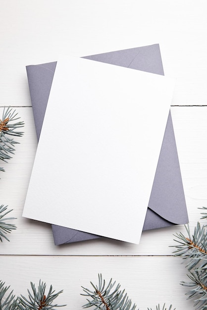 Maquete de cartão de saudação de Natal com envelope cinza e galho de árvore de abeto verde fresco sobre fundo branco de madeira, vista superior plana, cartão de férias de inverno vazio
