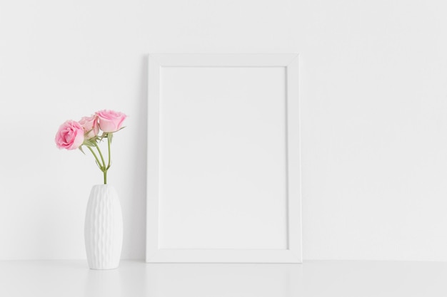 Maquete de moldura branca com rosas cor de rosa em um vaso em uma mesa brancaOrientação de retrato